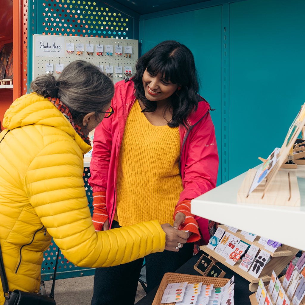 Nerg and a customer interacting at an outdoor market stall with colourful jewellery products in the background.