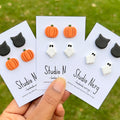 Three sets of Autumn and Halloween-themed stud earrings (black cats, orange pumpkins and white ghosts) on cards held by a hand with a grassy background.