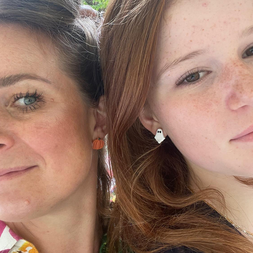Close-up of two women wearing Autumn themed earrings - one with handmade orange pumpkin stud earrings and the other wearing handmade white ghost stud earrings.