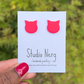Handmade hot pink cat-shaped earrings on a 'Studio Nerg' card against a blurred green nature background.