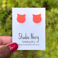Handmade orange cat-shaped earrings on a 'Studio Nerg' card against a blurred green nature background.