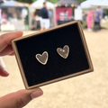 Handmade heart-shaped ceramic stud earrings with distressed brown colouring in a gift box with luxury black velvet, held by a hand with a blurred outdoor background.