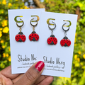 Two pairs of handmade ladybird charms on gold and silver huggie hoop earrings on 'Studio Nerg' branded cards held by a hand against a blurred nature background.
