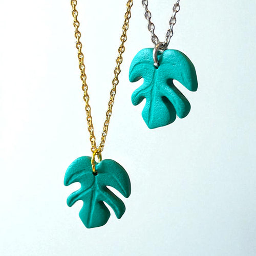 Two necklaces with a handmade green monstera leaf pendants on gold and silver chain with a white background.