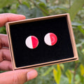 Handmade-round ceramic stud earrings with a split red and white design in a gift box with luxury black velvet, held by a hand with a blurred outdoor background.