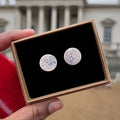 Handmade round ceramic stud earrings in white with blue speckle design in a gift box with luxury black velvet, held by a hand at a low angle with a blurred outdoor background.