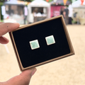 Handmade square ceramic stud earrings with pale mint blue colouring with  speckles in a gift box with luxury black velvet, held by a hand with a blurred outdoor background.