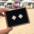 Handmade square ceramic stud earrings in white with black speckles in a gift box with luxury black velvet, held by a hand with a blurred outdoor background.