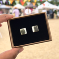 Handmade square ceramic stud earrings with split white and grey metallic glaze effect in a gift box with luxury black velvet, held by a hand with a blurred outdoor background.