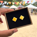 Handmade square yellow ceramic stud earrings in a gift box with luxury black velvet, held by a hand with a blurred outdoor background.