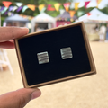 Handmade square ceramic stud earrings with a striped distressed teal colouring in a gift box with luxury black velvet, held by a hand with a blurred outdoor background.