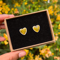 Handmade heart-shaped ceramic stud earrings with a crackled yellow and black glaze effect in a gift box with luxury black velvet, held by a hand with a blurred nature background.