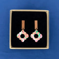A pair of dangling earrings displayed in a square kraft presentation box on a black velvet insert, set against a solid blue background. The earrings feature a colourful marbled pattern in pink, green, orange on white, suspended from a natural wood bar.