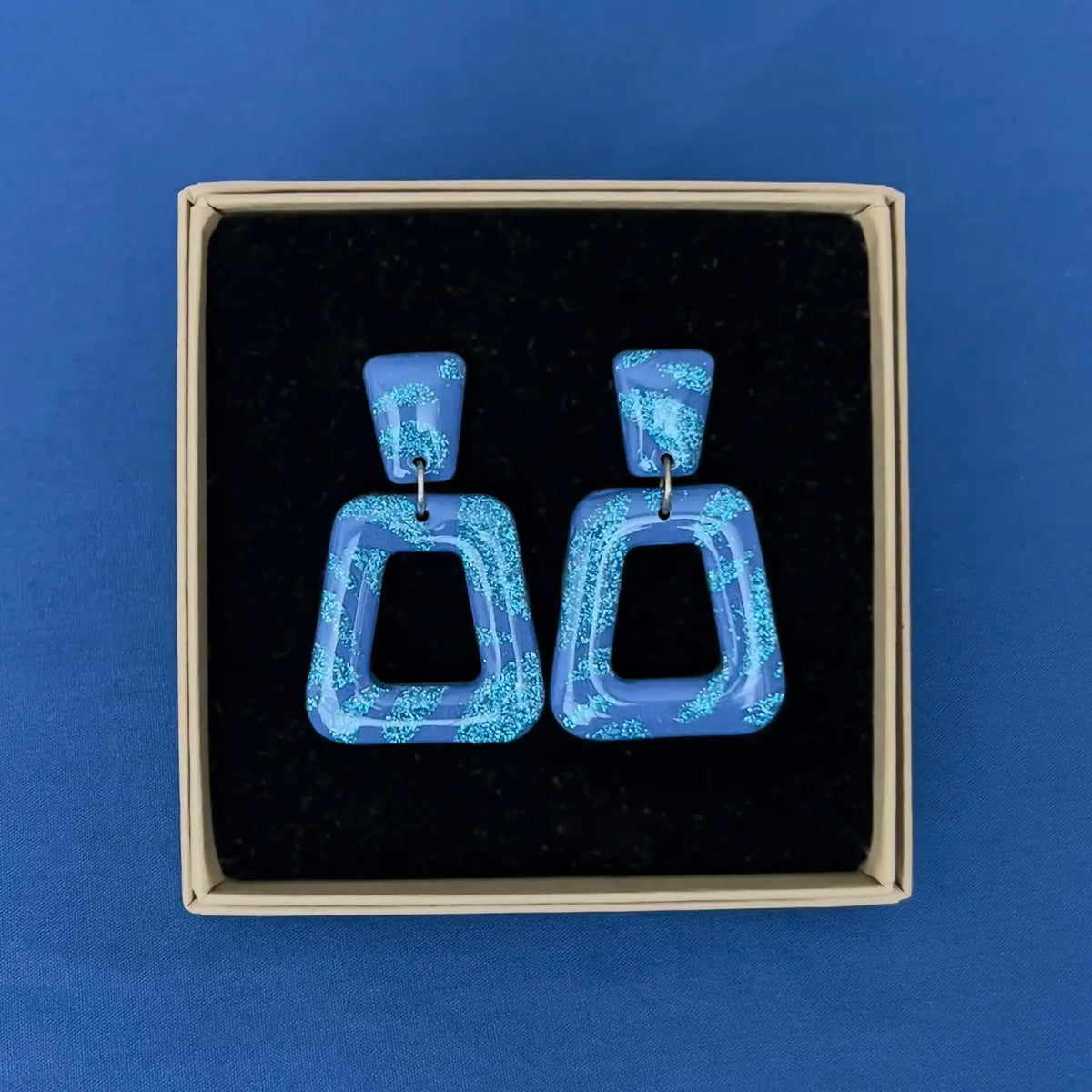 A pair of deep blue geometric drop earrings with a glittery light blue pattern, presented in a kraft jewellery box with black velvet insert on a solid blue background.
